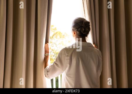 Rideaux d'ouverture, femme mature en chemise blanche regardant à l'extérieur de la fenêtre à la maison, espace de copie Banque D'Images