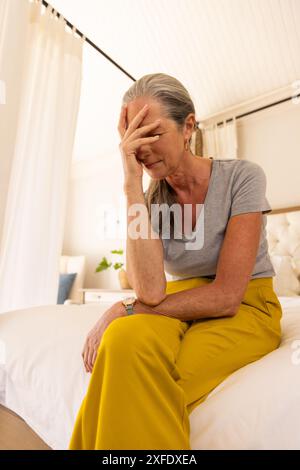 Assise sur le lit, femme mature en pantalon jaune paraissant stressée et fatiguée Banque D'Images