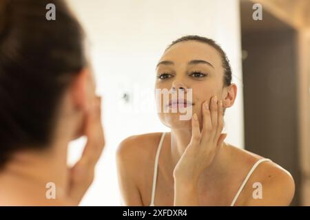 Jeune femme examinant son visage dans le miroir, se concentrant sur la routine de soin de la peau Banque D'Images