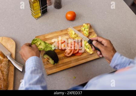 Préparation des toasts d'avocat et de tomate, homme tranchant les légumes sur une planche à découper en bois Banque D'Images