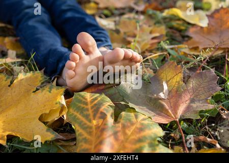 enfant pieds nus assis sur le sol parmi les feuilles d'automne jaunes tombées dans le parc. mignons orteils d'enfants illuminés par le soleil d'automne, la joie, l'atmosphère positive, hap Banque D'Images