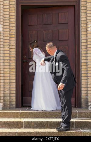 La mariée et le marié sont debout sur les marches contre la toile de fond des portes brunes. Le marié garde ses mains dans ses poches. La mariée se tient derrière t Banque D'Images