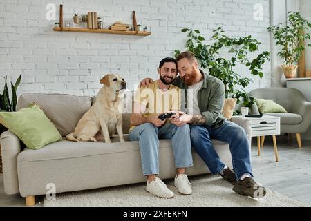 Deux hommes, un couple gay, assis sur un canapé en jouant à un jeu vidéo avec leur chien labrador dans un cadre confortable de salon. Banque D'Images