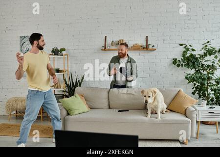 Deux hommes barbus jouant avec leur chien Labrador dans un salon confortable tout en regardant un match sportif à la télévision. Banque D'Images