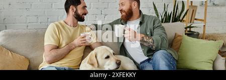 Couple gay barbu assis sur le canapé avec chien Labrador tout en tenant des tasses de café dans le salon confortable. Banque D'Images