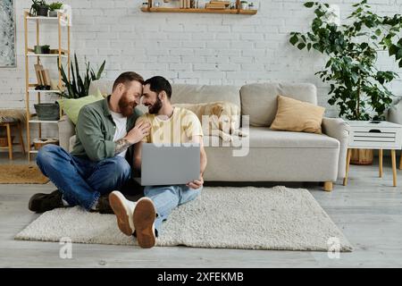 Couple gay barbu travaillant à distance sur le sol avec un ordinateur portable, accompagné de leur fidèle chien labrador. Banque D'Images