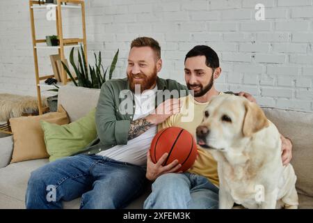 Couple gay barbu profitant de temps de qualité sur le canapé avec leur chien Labrador et un basketball. Banque D'Images