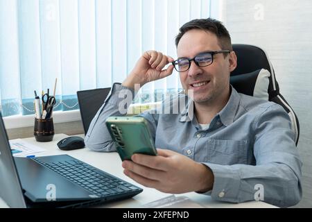 Bel homme d'affaires utilise un smartphone et sourit tout en travaillant au bureau. un employé masculin regarde une vidéo sur un smartphone. Banque D'Images
