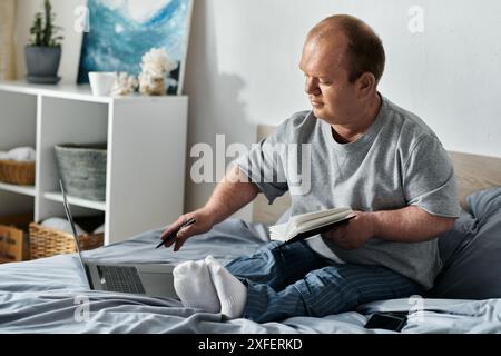 Un homme inclusif est assis sur son lit, travaillant sur un ordinateur portable et lisant un livre. Banque D'Images