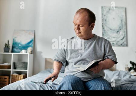 Un homme inclusif est assis sur le bord de son lit, lisant un livre et regardant son téléphone. Banque D'Images