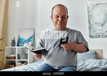 Un homme avec inclusivité est assis sur un lit, tenant un livre et vérifiant son téléphone. Banque D'Images