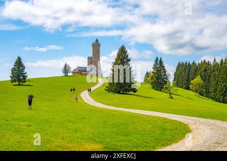 Un sentier de gravier sinueux mène à la tour d'observation de Dalimil en Tchéquie, entourée de champs verdoyants et d'arbres imposants. Le ciel est d'un bleu vif avec des nuages blancs moelleux. Banque D'Images