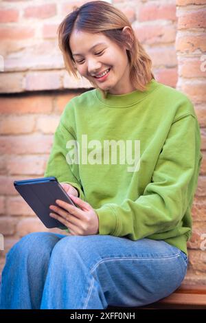 Jeune femme asiatique souriante assise surfant sur Internet sur une tablette numérique. Banque D'Images