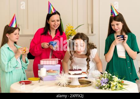 Une fille en robe blanche souffle des bougies d'anniversaire pendant que sa famille regarde et célèbre autour d'une table décorée. Banque D'Images