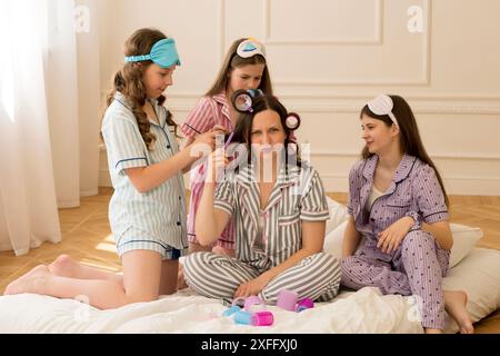 Une femme en pyjama rayé entourée de trois filles coiffant ses cheveux lors d’une nuit de nuit ludique, tout en souriant et en profitant du moment. Banque D'Images