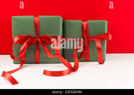 Deux cadeaux de Noël enveloppés dans du papier vert avec des noeuds en satin rouge sur une table blanche avec un fond rouge. Vue de face Banque D'Images