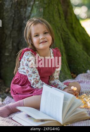 Une petite fille de 5 ans est assise près d'un grand arbre dans le parc. Sourit et regarde la caméra. A proximité se trouve un livre ouvert et des lanternes. Banque D'Images