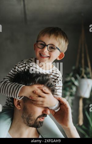 Portrait de garçon heureux couvrant les yeux du père tout en étant assis sur l'épaule Banque D'Images