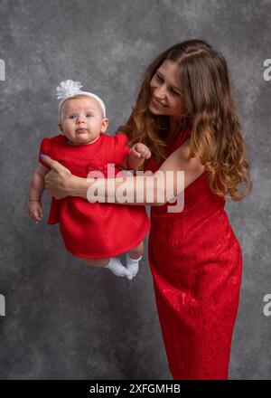 Maman tient sa petite fille dans ses bras. Tous deux portent des robes rouges. Tournage en studio. Tendresse et amour de mère. Liens familiaux Banque D'Images