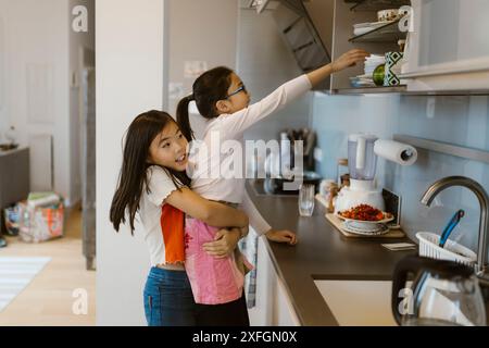 Vue latérale de la fille aidant la sœur à atteindre les armoires dans la cuisine Banque D'Images