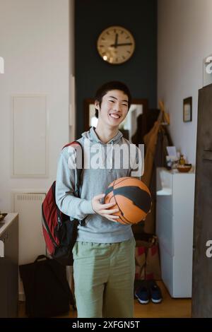 Garçon souriant avec sac à dos tenant le basket debout à la maison Banque D'Images