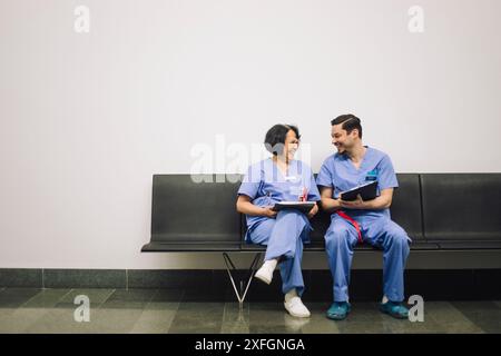 Heureux travailleurs médicaux hommes et femmes discutant tout en étant assis sur des chaises près du mur à l'hôpital Banque D'Images