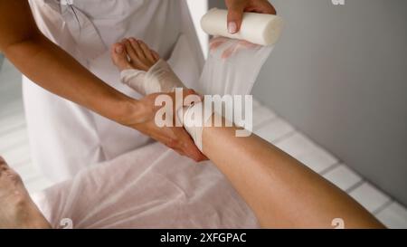 Le cosmétologue fait un bandage sur la jambe. Enveloppement corporel. Enveloppements brûlant les graisses. Procédure d'enveloppement et de bandage anti-cellulite. Une esthéticienne enveloppe la jambe du client. Banque D'Images