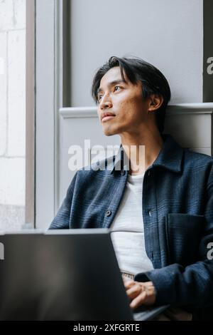 Homme d'affaires réfléchi regardant loin tout en étant assis avec ordinateur portable près de la fenêtre au bureau Banque D'Images