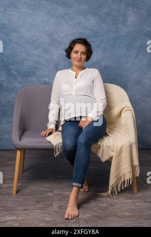 Femme heureuse enceinte en jeans et une blouse blanche se trouve sur le canapé. Portrait d'une femme enceinte d'âge moyen. Concept de grossesse saine, brune wom Banque D'Images