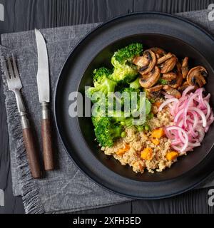 quinoa à la patate douce, champignons sautés, oignon rouge mariné, pois verts, brocoli bouilli dans un bol blanc sur une table en bois noir avec couverts, coriandre, Banque D'Images