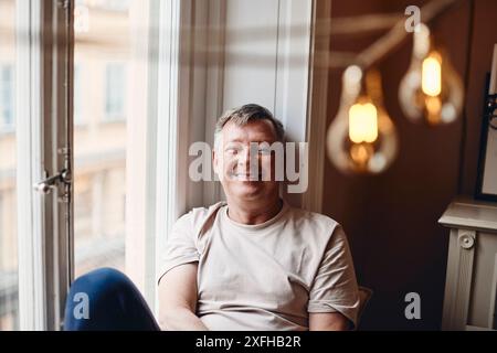 Portrait d'homme mature souriant assis près de la fenêtre à la maison Banque D'Images