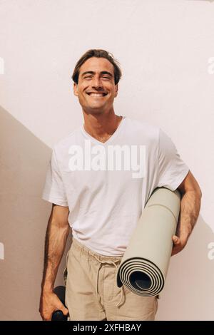 Portrait d'homme souriant tenant un tapis de yoga enroulé et debout près du mur Banque D'Images