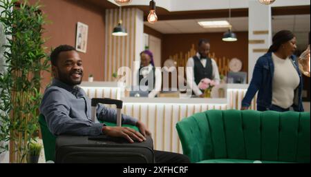 Client de l'hôtel attendant à la réception pour l'enregistrement, assis dans le salon à la réception à côté des bagages et se préparant à voir la chambre. Homme afro-américain voyageant en vacances, arrivant à un complexe cinq étoiles. Banque D'Images