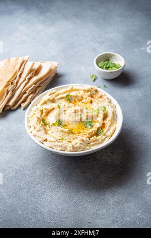 Bol de houmous crémeux garni d'huile d'olive et d'herbes est servi avec du pain pita sur une planche de bois. Un petit bol de garniture est placé à côté du Banque D'Images