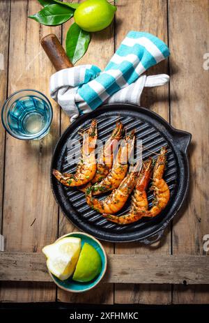 Crevettes grillées sur une poêle grillée noire sur une table en bois rustique, avec des citrons verts, un tissu rayé bleu Banque D'Images