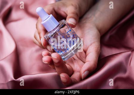 Flacon pipette en verre transparent avec sérum violet pour le visage, soins du corps dans les mains sur textile rose soyeux. Soins de beauté féminins. Produit esthéticien Banque D'Images