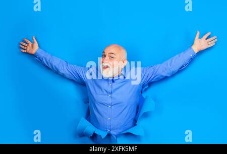 Souriant vieil homme barbu avec des lentilles de contact bleues regardant à travers le trou dans le papier. Heureux homme senior chauve dans la chemise élégante a levé les mains. Vente et remise Banque D'Images