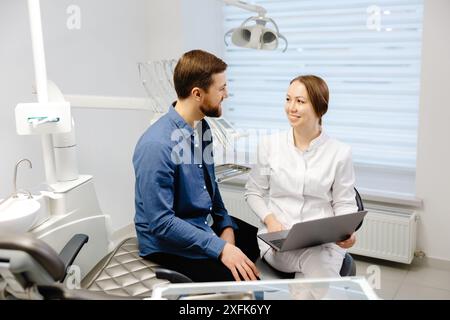 Un beau jeune homme parle à une femme médecin à un rendez-vous dentaire dans un beau bureau lumineux. Le dentiste explique au patient et montre Banque D'Images