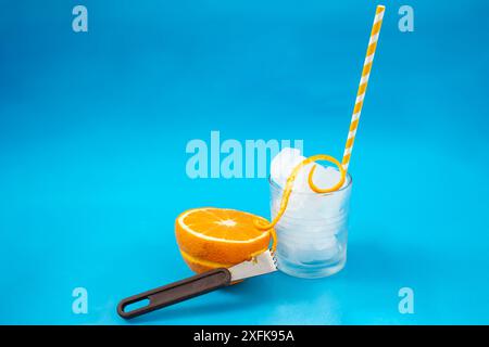 Citrus Zester, un demi-cube de fruit orange et un verre rempli de glace sur fond bleu, flou Banque D'Images
