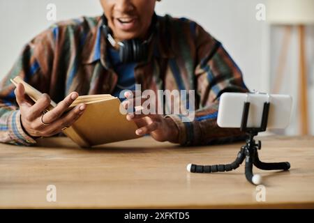 Un jeune homme afro-américain lit un livre tout en conversant sur la caméra de son téléphone à une table. Banque D'Images