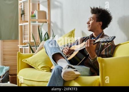 Un jeune afro-américain joue de la guitare acoustique assis sur un canapé jaune. Banque D'Images