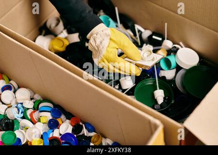 Une boîte déborde de gobelets de différentes couleurs, triés par un jeune volontaire en gants et gilet de sécurité pour le recyclage. Banque D'Images