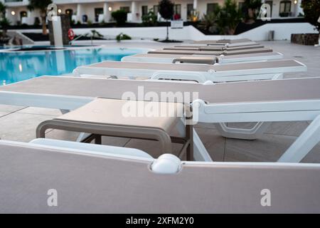 Une rangée de chaises longues est alignée sur la terrasse de la piscine. Les chaises sont blanches et bronzées, et elles sont toutes face à la piscine. La scène dégage une ambiance détendue Banque D'Images