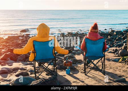 Deux personnes sur des chaises de plage ensemble sur la plage d'hiver, faire du thé chaud, du café le jour du ciel bleu ensoleillé et la mer en arrière-plan. Hiver froid britannique. La vie Banque D'Images