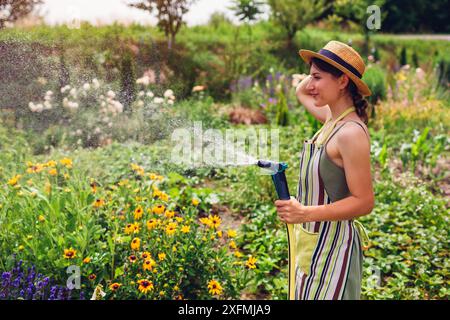 Jardinier de femme joyeuse dans le chapeau de paille tenant le tuyau de jardin ayant le plaisir d'arroser les plantes dans le jardin d'été. Prendre soin des fleurs à la ferme Banque D'Images