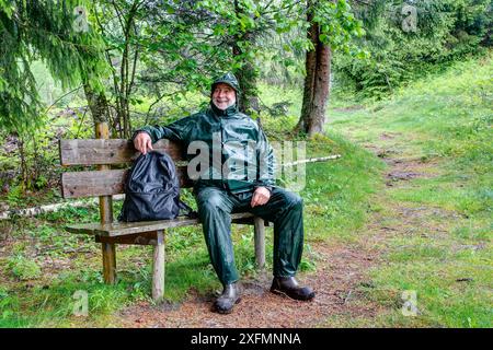 Sur le chemin du 'Kreuzboden Hut', un randonneur plus âgé fait une pause sur un banc le long du sentier de randonnée et est de bonne humeur malgré le temps pluvieux. Banque D'Images