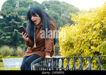 Surprise jeune femme regardant les notifications des médias sociaux au téléphone, à l'extérieur assis dans le parc passer l'après-midi, se reposer, copier l'espace Banque D'Images