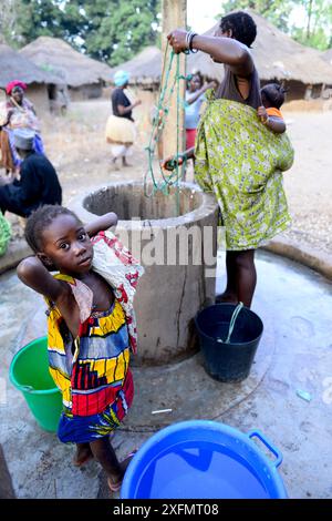 Mère avec bébé sur le dos recueillant de l'eau du puits, fille regardant la caméra au premier plan, île de Formosa, réserve de biosphère de l'UNESCO de Bijagos, Guinée Bissau, février 2015. Banque D'Images