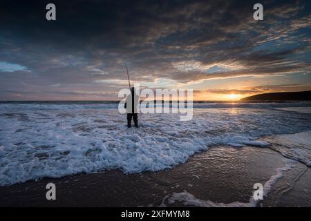 Pêcheur à la ligne au crépuscule dans le surf de mer. L'achigan (Dicentrarchus labrax) est souvent attrapé alors qu'il chasse à la nourriture barbée par les vagues, Porth Ceiriad, Pwllheli, Gwynedd, pays de Galles, Royaume-Uni, décembre 2016. Banque D'Images