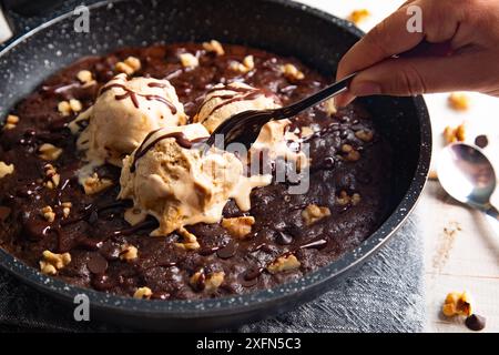 Brownie fait maison cuit dans une poêle, non cuit, avec des cuillères de crème glacée à la vanille fondante, des noix et des pépites de chocolat et une main de femmes prenant un morceau d'esprit Banque D'Images